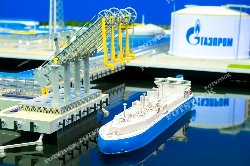 На фотографии изображён макет терминала для перевалки сжиженного газа с судном у причала. Детализированная модель иллюстрирует промышленную инфраструктуру и технологии транспортировки газа. На заднем плане виден логотип компании, связанной с газовой промышленностью.